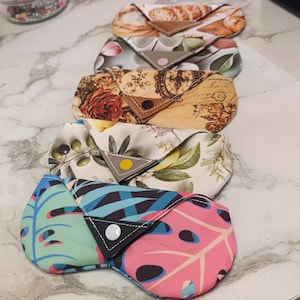 May include: Five envelope-style fabric eyeglass cases arranged in a vertical row. Each case features a unique print, including bread, floral patterns, vintage script, olives with lemons, and vibrant tropical leaves with pink and blue tones.