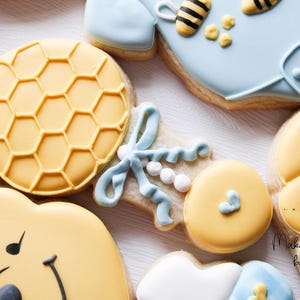 May include: A set of decorated sugar cookies featuring a honeybee theme. The cookies include a baby onesie, a rattle, a bear, and a honey comb. The cookies are decorated with yellow, blue, and white icing.