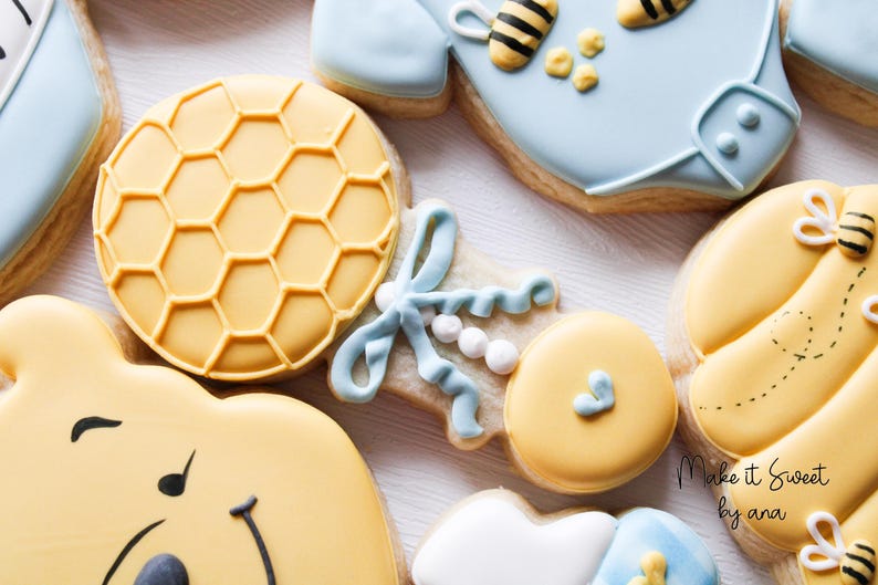 May include: A set of decorated sugar cookies featuring a honeybee theme. The cookies include a baby onesie, a rattle, a bear, and a honey comb. The cookies are decorated with yellow, blue, and white icing.