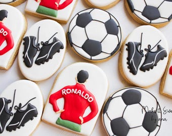 Puede incluir: Galletas de azúcar decoradas con temática de fútbol. Las galletas muestran un balón de fútbol, botas de fútbol y un jugador con una camiseta roja con el nombre "RONALDO" en la espalda.