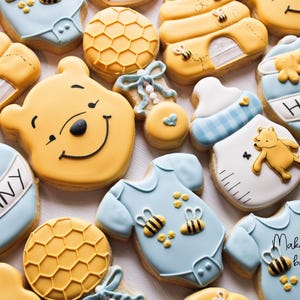 May include: A collection of Winnie the Pooh themed sugar cookies decorated with blue, yellow, and white icing. The cookies feature images of Winnie the Pooh, honey pots, beehives, and baby onesies. The cookies are perfect for a baby shower or birthday party.