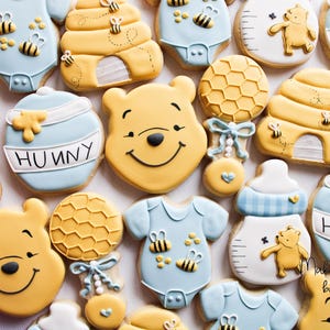 May include: A set of Winnie the Pooh themed sugar cookies decorated with blue, yellow, and white icing. The cookies feature Winnie the Pooh's face, a honey pot, a beehive, a rattle, and a baby onesie. The cookies are decorated with the word "Hunny".