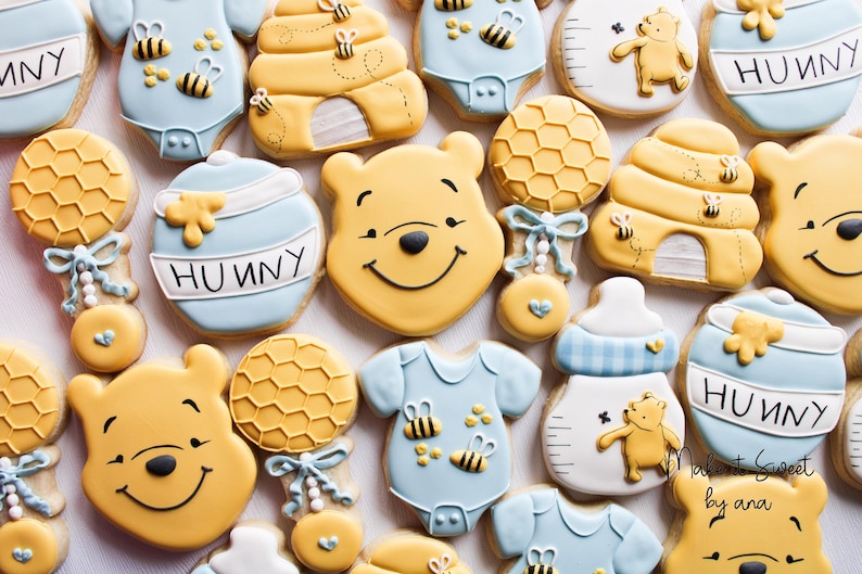 May include: A set of Winnie the Pooh themed sugar cookies decorated with blue, yellow, and white icing. The cookies feature Winnie the Pooh's face, a honey pot, a beehive, a rattle, and a baby onesie. The cookies are decorated with the word "Hunny".