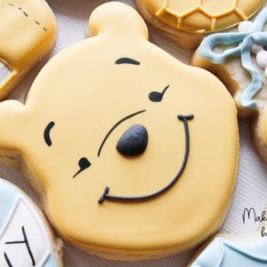 May include: A yellow sugar cookie decorated with black icing to look like Winnie the Pooh. The cookie has a smiling face with a black nose and a black outline around the mouth.