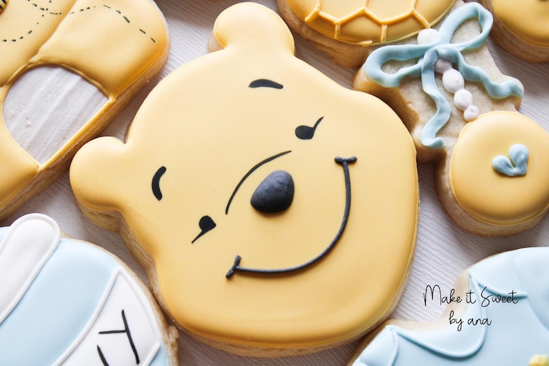 May include: A yellow sugar cookie decorated with black icing to look like Winnie the Pooh. The cookie has a smiling face with a black nose and a black outline around the mouth.