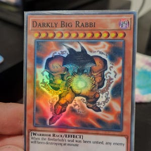 May include: A holographic trading card with the name "Darkly Big Rabbi" displayed at the top. The card features a monster illustration and text that reads "WARRIOR RACE/EFFECT" and "ATK/INFINITY DEF/INFINITY".