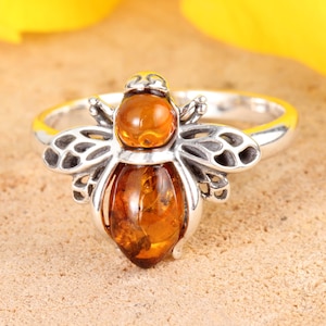 Peut inclure: Bague en argent ornée d'une abeille. Le corps et la tête de l'abeille sont en pierre de couleur ambre, avec des ailes argentées détaillées. La bague est présentée sur un fond neutre, mettant en valeur le savoir-faire de la bijouterie.