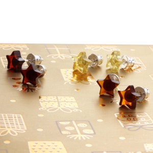 May include: Five pairs of star-shaped amber earrings in various shades of brown and yellow. The earrings are displayed on a gold background with a gift wrap pattern.