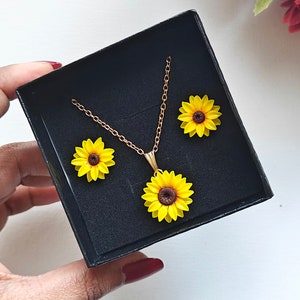 May include: A gold chain necklace with a sunflower pendant and two matching sunflower earrings. The jewelry is displayed in a black velvet box.
