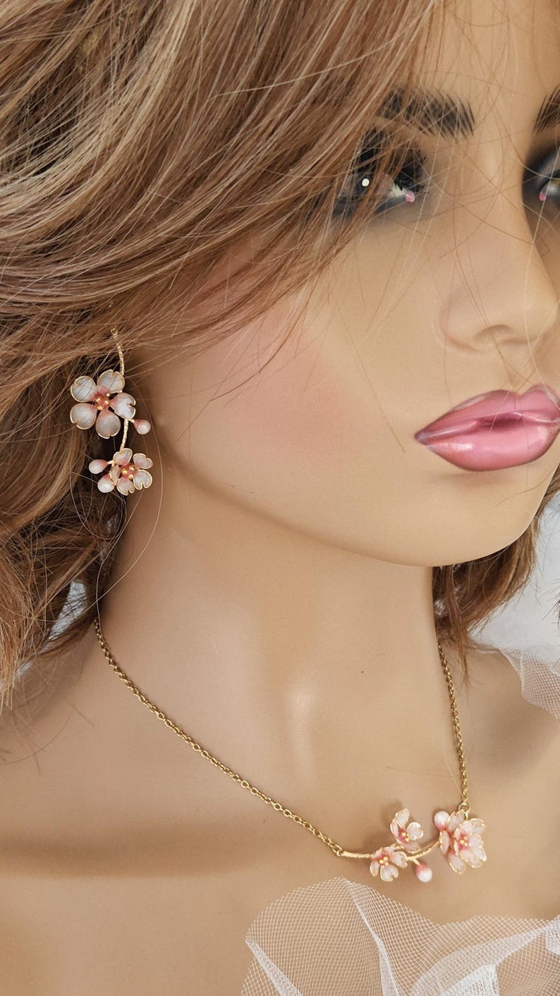 May include: Delicate floral jewelry set featuring a gold-tone necklace and earrings. The necklace and earrings are adorned with clusters of pink and white flower-shaped pendants. The earrings hang from a gold wire, and the necklace rests on the collarbone.