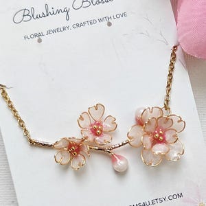 May include: A delicate gold-toned necklace featuring three pink and white floral blossoms. The blossoms are detailed with pink centers and a pearlescent drop. The card reads "Blushing Blossoms Floral Jewelry, Crafted with Love."