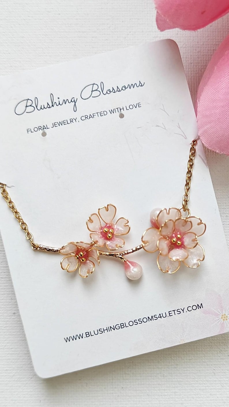 May include: A delicate gold-toned necklace featuring three pink and white floral blossoms. The blossoms are detailed with pink centers and a pearlescent drop. The card reads "Blushing Blossoms Floral Jewelry, Crafted with Love."