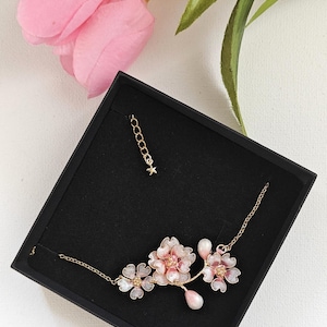 May include: A delicate floral necklace with pink and white blossoms, presented in a black box. The necklace features a gold chain and a small star charm. Pink flowers and green leaves are in the background.