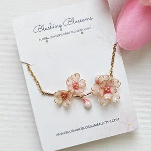 May include: A delicate floral necklace featuring pink and white blossoms with gold-colored accents. The necklace is displayed on a white card with the text "Blushing Blossoms" and "Floral Jewelry, Crafted with Love."