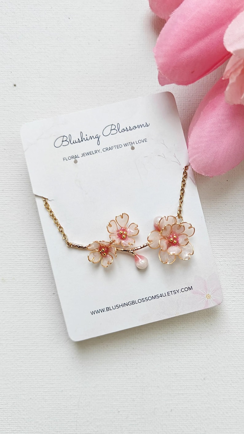 May include: A delicate floral necklace featuring pink and white blossoms with gold-colored accents. The necklace is displayed on a white card with the text "Blushing Blossoms" and "Floral Jewelry, Crafted with Love."