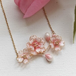 May include: A delicate gold-tone necklace featuring a cluster of pink cherry blossom flowers with white accents and pear-shaped pendants. The floral design is set against a soft pink background.