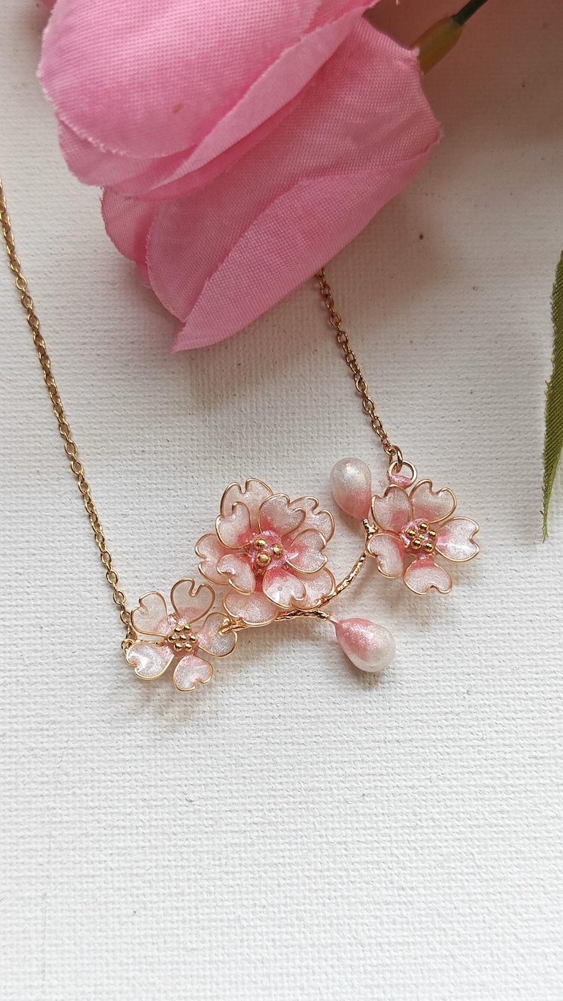 May include: A delicate gold-tone necklace featuring a cluster of pink cherry blossom flowers with white accents and pear-shaped pendants. The floral design is set against a soft pink background.
