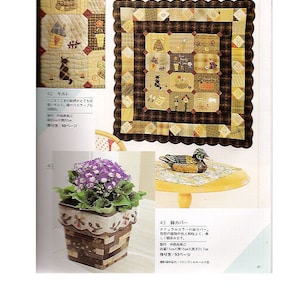 May include: A close-up of a brown and beige patchwork quilt with a scalloped border. The quilt is made up of squares with different patterns and designs, including a cat, a house, and a beehive. The quilt is displayed on a table with a wooden duck figurine and a potted plant with purple flowers.