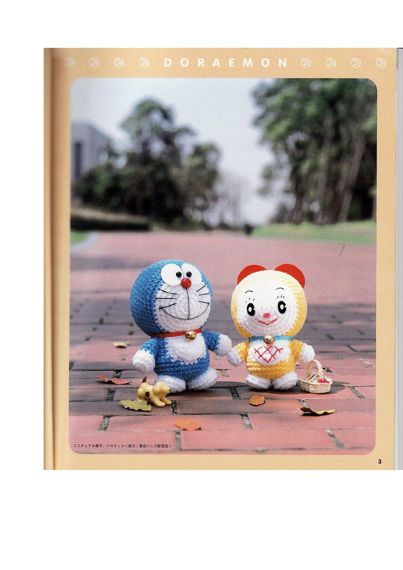 May include: Two crocheted stuffed toys, one blue and one yellow, standing on a brick pathway. The blue toy is a character from the anime Doraemon, and the yellow toy is a smaller character with a red bow. The toys are surrounded by fallen leaves.