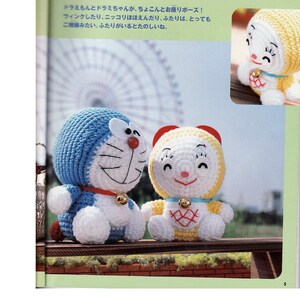 May include: Two crocheted stuffed toys, one blue and white and one yellow and white, are sitting on a wooden bench. The blue and white toy is a cat with a red nose and a bell around its neck. The yellow and white toy is a cat with a pink nose and a bell around its neck. The toys are smiling and have their eyes closed. The background is a blurry image of a Ferris wheel.