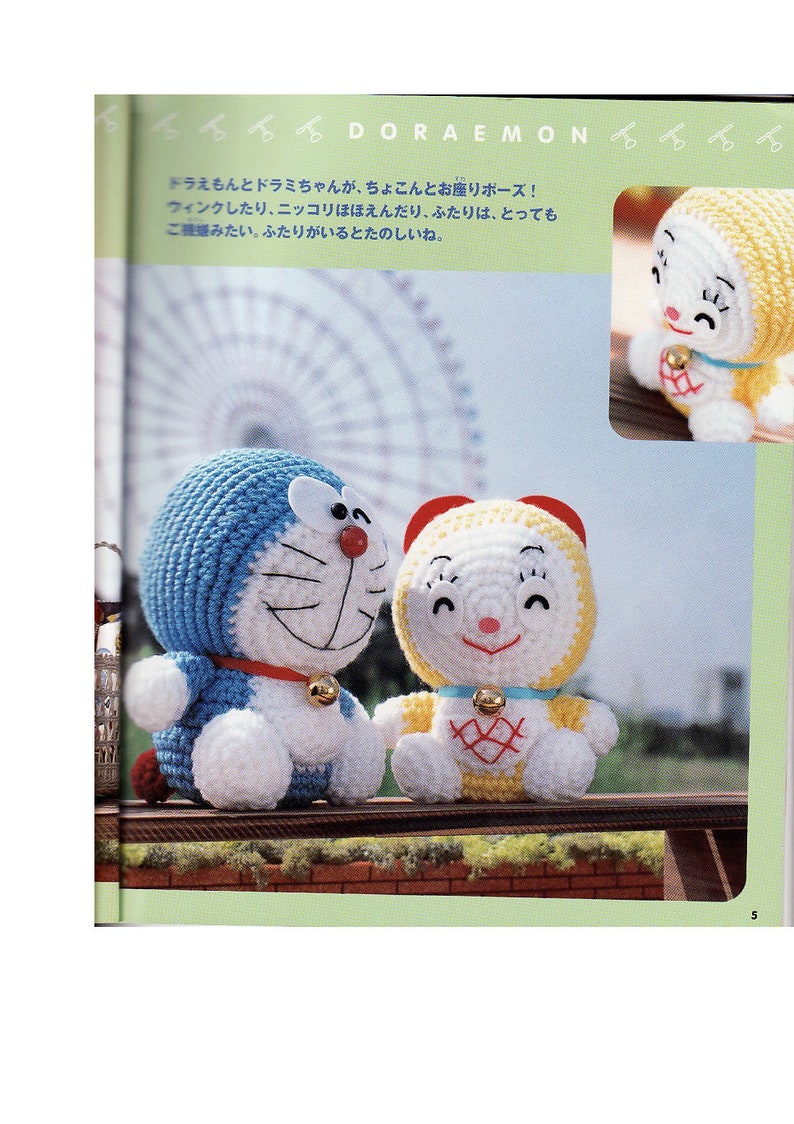 May include: Two crocheted stuffed toys, one blue and white and one yellow and white, are sitting on a wooden bench. The blue and white toy is a cat with a red nose and a bell around its neck. The yellow and white toy is a cat with a pink nose and a bell around its neck. The toys are smiling and have their eyes closed. The background is a blurry image of a Ferris wheel.