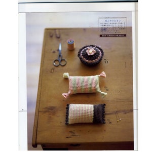 Peut inclure: Trois coussins à épingles en crochet sur une surface en bois. Les coussins à épingles sont en forme d'oreillers et sont décorés de rayures roses et vertes, de rayures brunes et roses et d'un gâteau brun et blanc avec une fleur rose sur le dessus. Les coussins à épingles sont numérotés 12, 13 et 14.