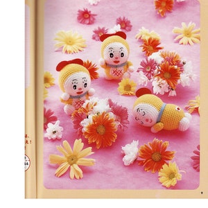 May include: Three crocheted dolls with yellow bodies, white faces, and red bows. The dolls are surrounded by pink and white flowers on a pink background. The text "DORAEMON" is printed in the top center of the image.