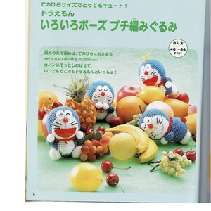 May include: Four crocheted Doraemon plushies in different poses, surrounded by fruit, including bananas, cherries, grapes, lemons, apples, and a pineapple. The plushies are approximately 12 inches tall. The text on the page reads "Doraemon" and "Various Poses Crochet Amigurumi."