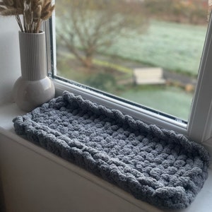 May include: A grey knitted cushion or throw blanket with a textured surface. The cushion is on a white windowsill.