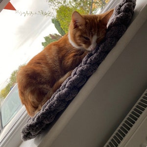 Pode incluir: Um gato ruivo com marcas brancas est&aacute; dormindo em uma cama de tric&ocirc; cinza. O gato est&aacute; enroscado na cama, que est&aacute; colocada em um parapeito de janela.