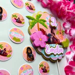 Op de afbeelding: Een verzameling kleurrijke feestversieringen met tropische thema's. Het middelpunt is een gelaagde taarttopper met een roze bloem, palmboom en de naam "Emilia". Extra ronde stickers hebben vergelijkbare ontwerpen en de naam "Emilia".