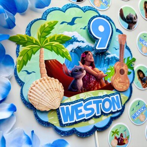 Op de afbeelding: Een kleurrijke taarttopper met een tropisch thema. Het heeft een palmboom, een zeeschelp en een cartoonafbeelding van een meisje en een blauwe alien. Het nummer "9" wordt weergegeven, samen met de naam "Weston".