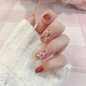 May include: Close-up of a hand with red and floral nail art. The nails are painted with a light pink base and have white flowers with black centres. Some nails have a gold accent.