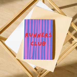 Könnte beinhalten: Eine Karte mit dem Text "RUNNERS CLUB" in Rot. Der Hintergrund ist lila mit vertikalen blauen und rosa Streifen. Die Karte liegt auf hellbraunem Papier, das sich auf weißem Papier in einem Holzrahmen befindet.
