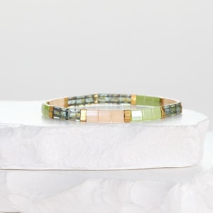 Puede incluir: Un brazalete de cuentas verdes, rosas y azules con detalles dorados. El brazalete está hecho de pequeñas cuentas cuadradas y está sobre una superficie blanca.