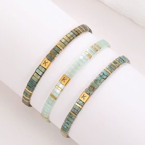 May include: Three bracelets with green and blue glass beads and gold accents. Each bracelet has a gold charm with the letter "K" engraved on it.