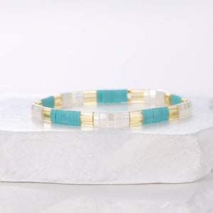 Peut inclure: Un bracelet en perles turquoise, blanc et or. Le bracelet est composé de petites perles carrées et est enfilé sur un élastique.