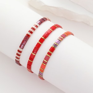 Peut inclure: Trois bracelets en perles colorées avec des perles rouges, blanches, orange et violettes. Les bracelets sont empilés les uns sur les autres sur une surface blanche.