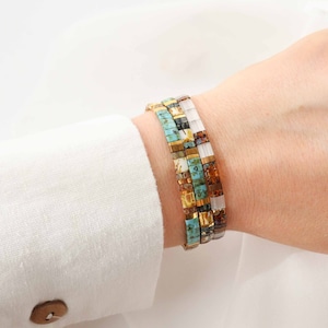 Puede incluir: Tres pulseras apiladas con cuentas cuadradas de colores. Las pulseras están hechas de cuentas turquesa, doradas, marrones y blancas.