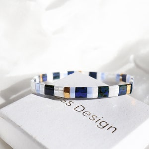 Könnte beinhalten: Eine weiße Box mit dem Aufdruck "Boss Design". Auf der Box liegt ein blaues, weißes und goldenes Armband.
