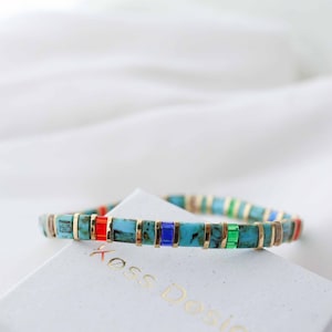 Puede incluir: Un brazalete de cuentas turquesa, dorado, rojo, verde y azul con un cierre dorado. El brazalete está sobre una caja blanca con el texto "Xoss Design" impreso en ella.