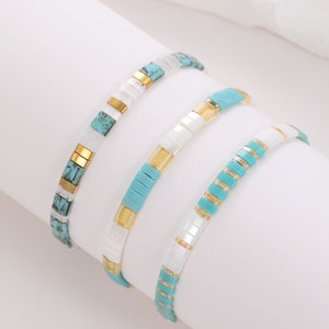 Peut inclure: Trois bracelets en perles avec des perles turquoise et blanches et des accents dorés. Les bracelets sont empilés les uns sur les autres.