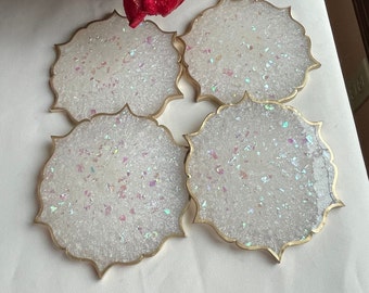 May include: Four white coasters with a gold rim and iridescent glitter embedded in the resin. The coasters are shaped like a flower with multiple points.