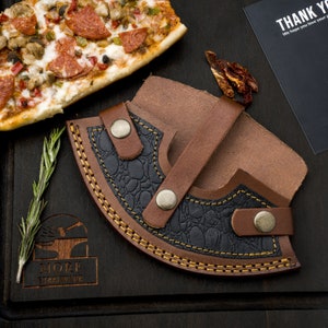 May include: A brown leather sheath with a black leather interior and a yellow stitching detail. The sheath has a strap and a snap closure. The sheath is on a wooden surface with a pizza slice and a rosemary sprig.