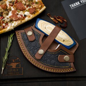May include: A blue and wood handled knife with a leather sheath. The sheath has a brown leather strap and a black leather lining. The knife is on a dark wooden surface.