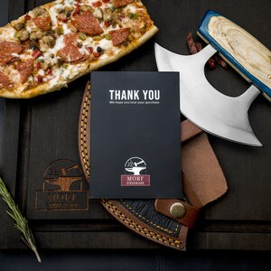 May include: A black card with the text "THANK YOU" and "We hope you love your purchase" on it. The card also has a logo of a blacksmith's anvil with a hammer on top of it and the text "MORF STEELWARE" below it. The card is sitting on a wooden cutting board with a pizza and a large knife with a leather sheath.