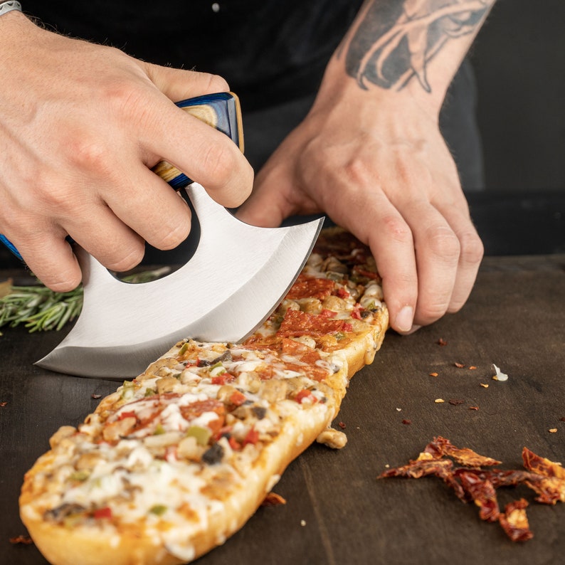 May include: A person uses a blue and silver handled knife to cut a long, rectangular pizza with pepperoni, mushrooms, and green peppers. The pizza is on a dark brown wooden surface.