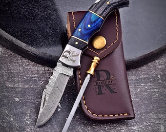May include: A folding pocket knife with a Damascus steel blade, a blue and black wood handle, and the name Richard engraved on the bolster. It is paired with a sharpening rod and a brown leather sheath embossed with the name Robert.