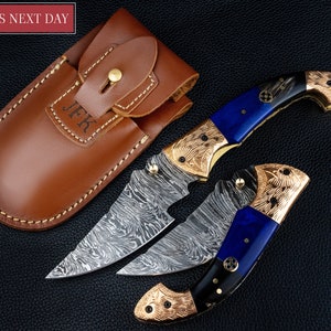 May include: Two Damascus steel knives with blue and black handles and gold accents. The knives are folded and have a decorative pattern on the blades. The knives come with a brown leather sheath.