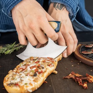 May include: A person uses a blue and brown handled knife to cut a long, rectangular pizza topped with pepperoni, mushrooms, green peppers, and cheese.